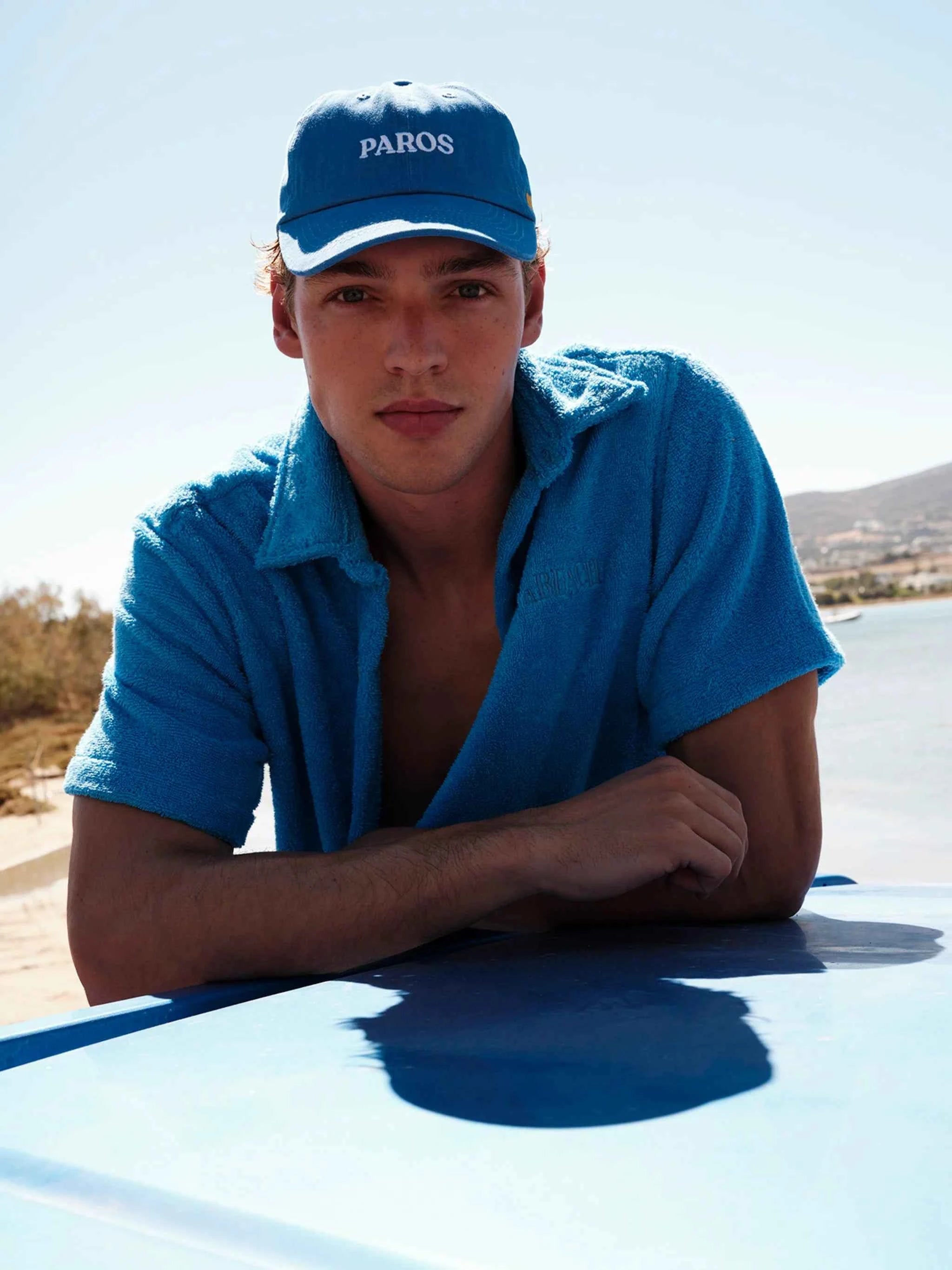 Man wearing a blue cap and shirt with 'PAROS' text, sitting on a car.