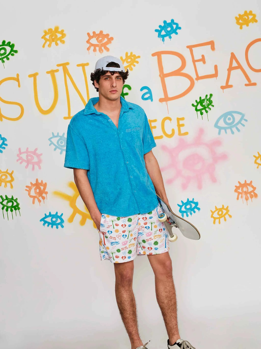 Person wearing a blue shirt and patterned shorts holding a skateboard against a colorful wall.