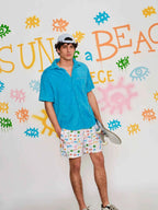 Person wearing a blue shirt and patterned shorts holding a skateboard against a colorful wall.