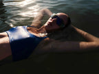 Person wearing a blue swimsuit and sunglasses, lying on their back in water.