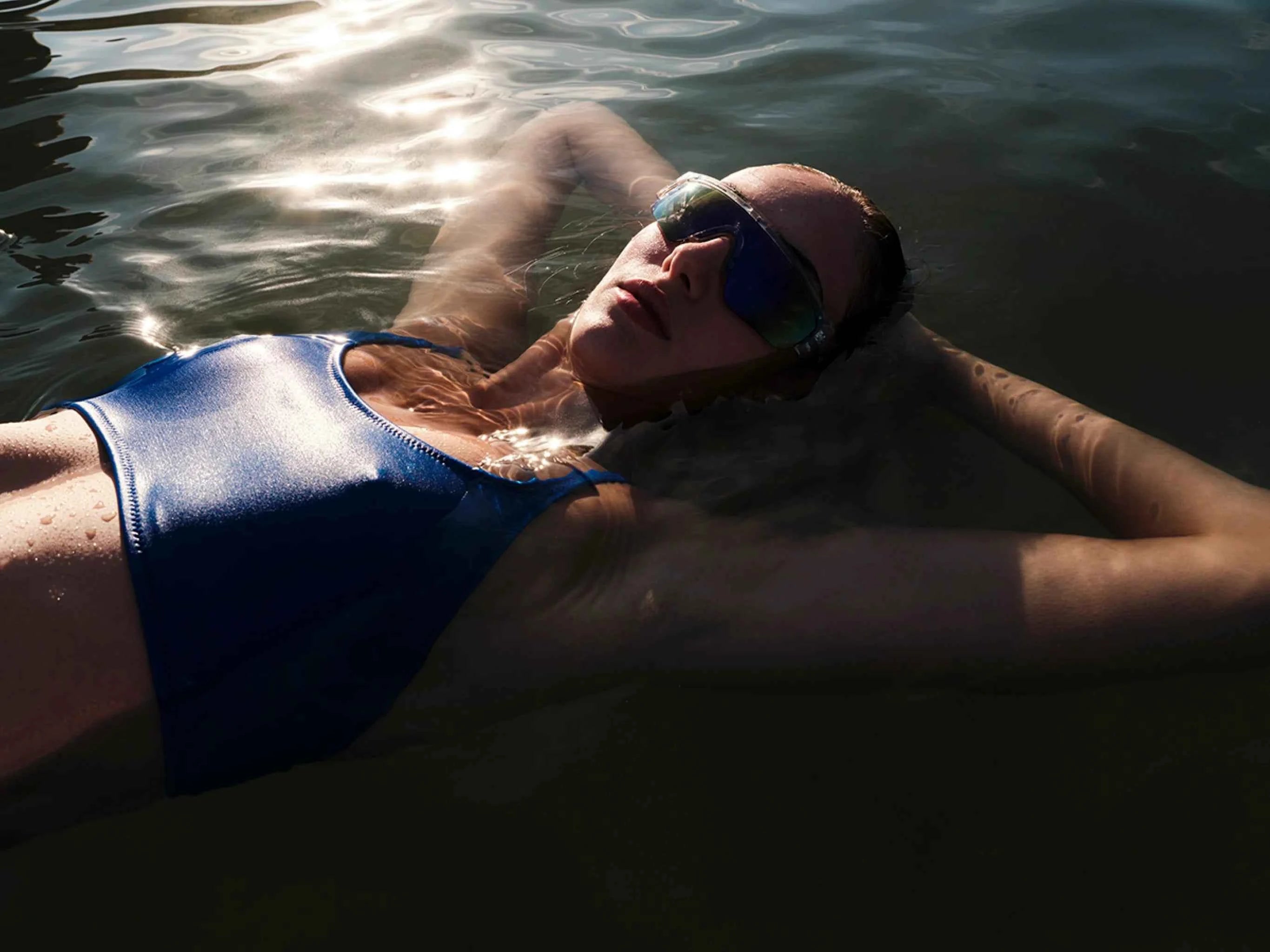 Person wearing a blue swimsuit and sunglasses, lying on their back in water.