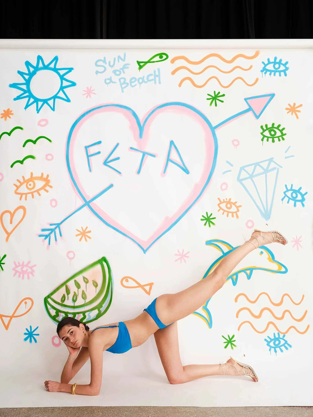 Person in blue bikini lying on a colorful beach-themed wall with 'FETA' written in a heart shape.