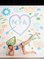 Person in blue bikini lying on a colorful beach-themed wall with 'FETA' written in a heart shape.