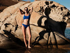 Woman in a blue bikini standing on a rocky beach with water in the background