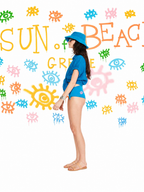 A woman stands in profile wearing a bright blue swim set, including a short-sleeve top and matching bikini-style bottoms, with a blue sun hat and sunglasses. The background is white with colorful painted suns, eyes, and the words “SUN OF BEACH GRECE.