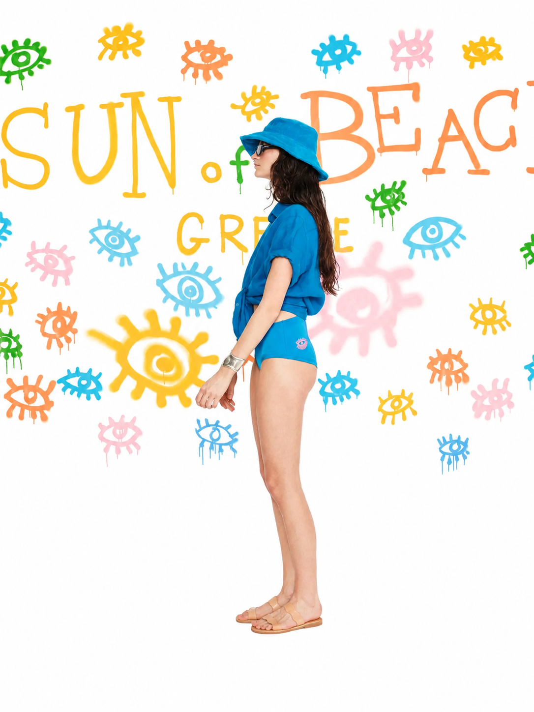A woman stands in profile wearing a bright blue swim set, including a short-sleeve top and matching bikini-style bottoms, with a blue sun hat and sunglasses. The background is white with colorful painted suns, eyes, and the words “SUN OF BEACH GRECE.