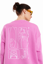 A woman shown from the back wearing a bright pink sweatshirt with white surf-themed graphic prints, including a palm tree, surfboard, jeep, and text reading 'SURF THE OFF'. She is wearing yellow-tinted sunglasses against a plain white background.