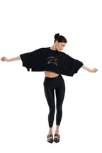 A woman modeling a black cropped tunic top with colorful text that reads 'FANTASY IS LIFETIME' paired with black leggings and black-and-white patterned shoes, set against a solid white background.