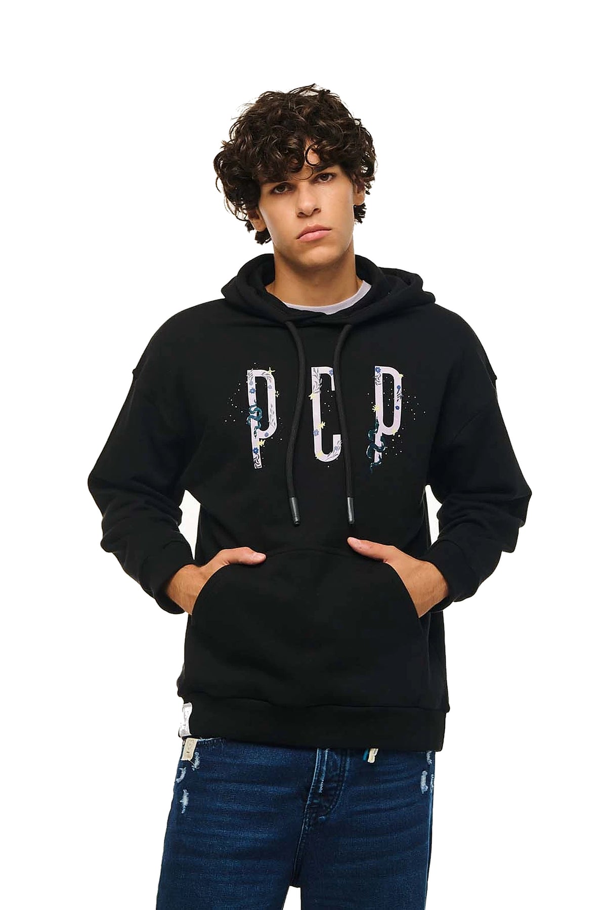 A young man with curly dark hair wearing a black pullover hoodie with 'PCP' printed in distressed white letters on the chest, paired with blue jeans, against a plain white background.