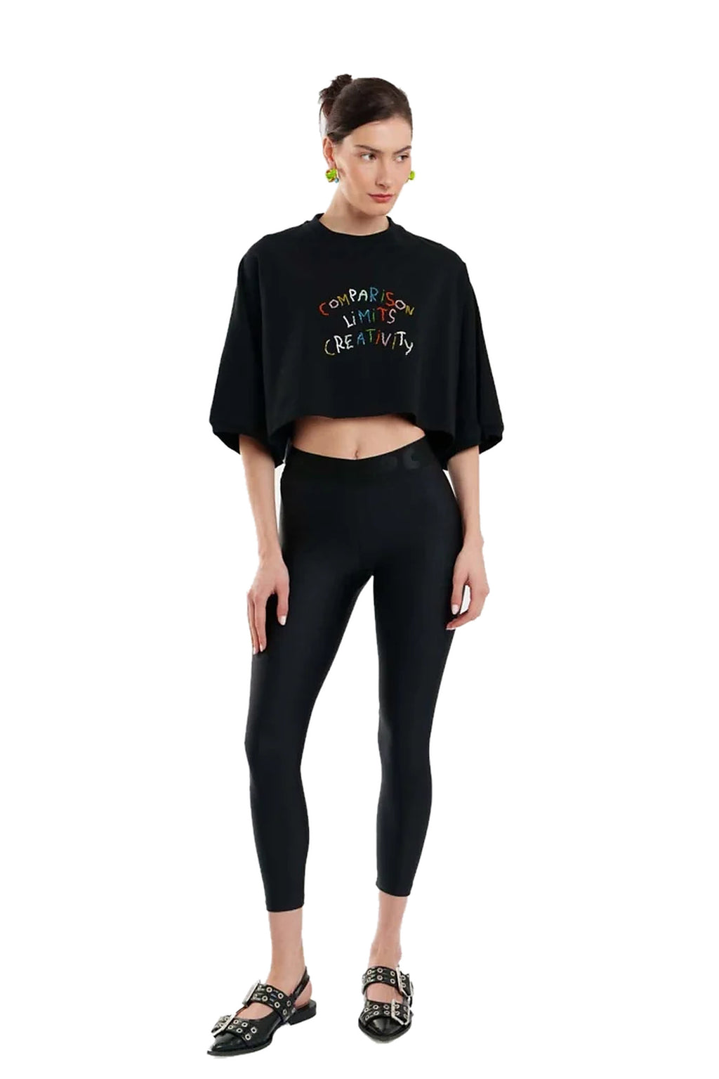 A model wearing a black cropped t-shirt with colorful text and black leggings, paired with black strappy flat shoes, against a plain white background.