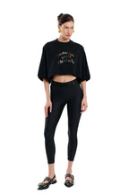 A model wearing a black cropped t-shirt with colorful text and black leggings, paired with black strappy flat shoes, against a plain white background.