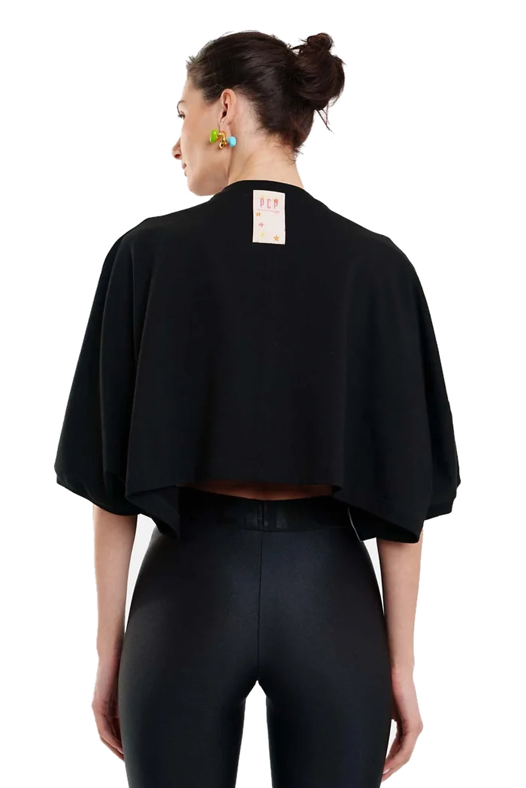 A woman shown from the back wearing a black cropped cape-style top and black leggings against a plain white studio background.