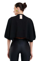 A woman shown from the back wearing a black cropped cape-style top and black leggings against a plain white studio background.