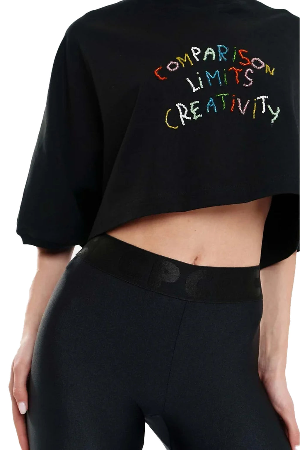 A cropped black t-shirt with the phrase 'COMPARISON LIMITS CREATIVITY' written across the chest in a colorful, hand-drawn multicolor font. The t-shirt is modeled with black leggings against a plain white background.