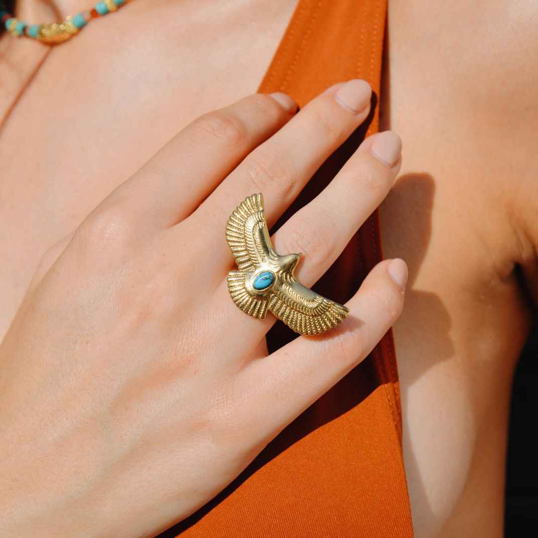 Gold ring with wings and turquoise stone on a hand against an orange background