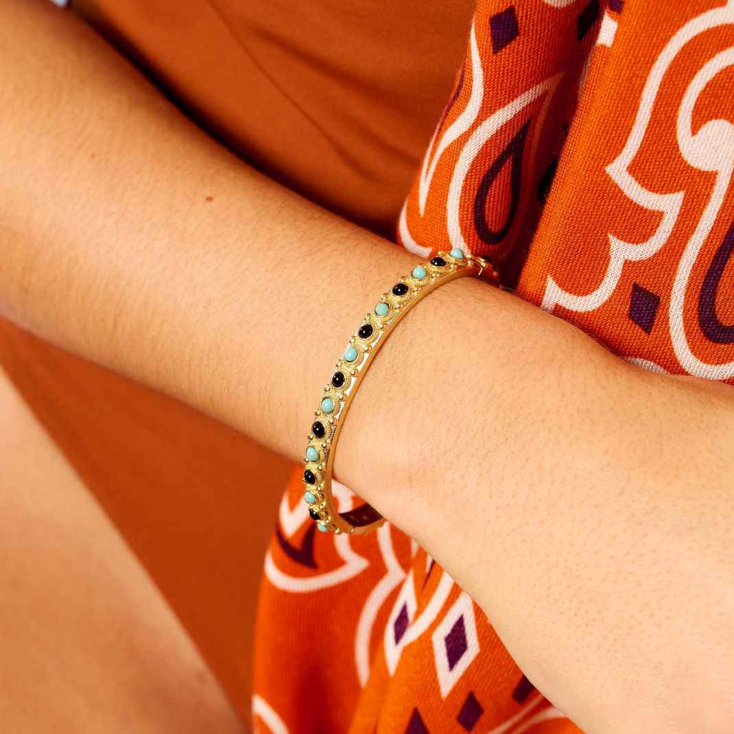Gold bracelet with turquoise and black stones on a wrist against an orange fabric background