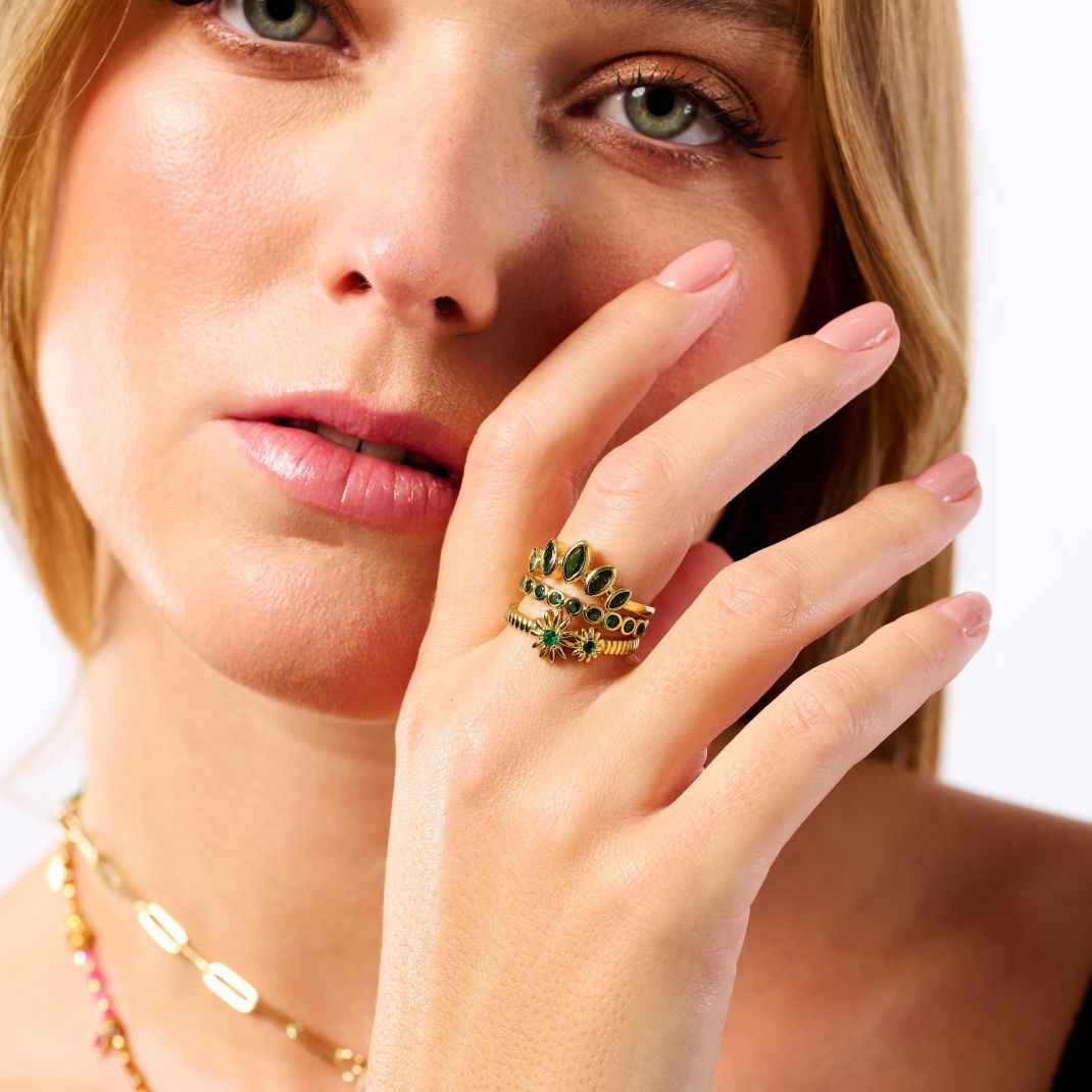 Close-up of a woman's hand wearing a gold ring with green gemstones, against a neutral background.