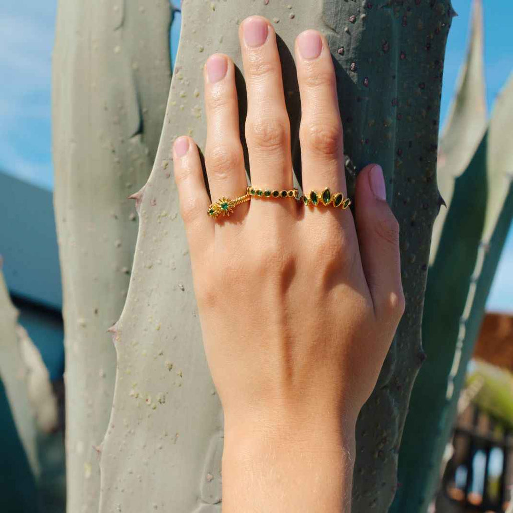 Hand wearing a gold ring with green gemstones against a cactus background