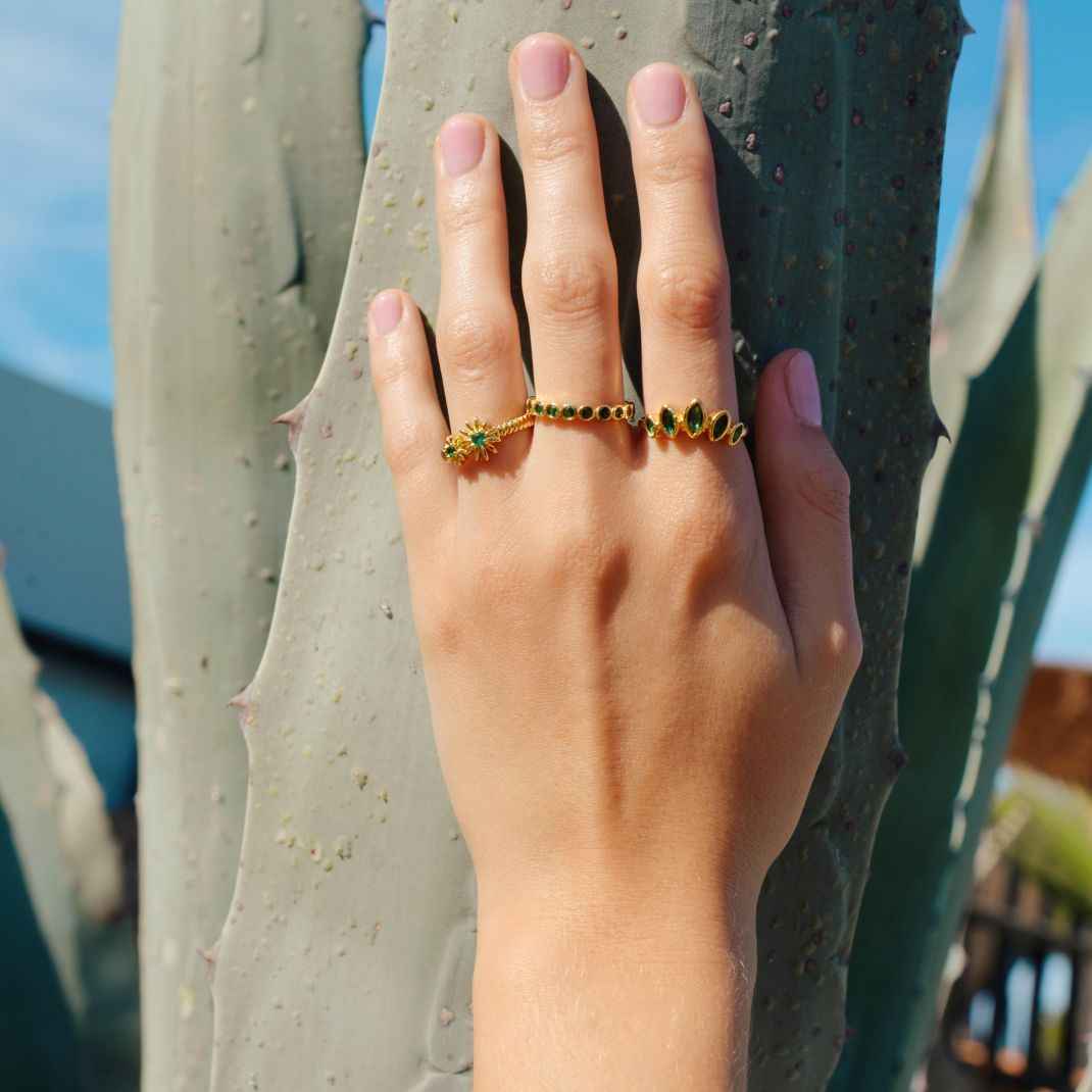 Hand wearing a gold ring with green gemstones against a cactus background