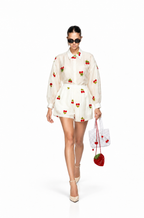 A model walks against a white studio background wearing a cream-colored two-piece set with red cherry and green leaf embroidery, paired with beige heels and holding a white strawberry-patterned handbag.