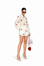 A model wearing a white long-sleeve shirt and matching shorts set, both decorated with red strawberry prints, styled with beige high heels, sunglasses, and a matching strawberry-patterned handbag against a plain white background.