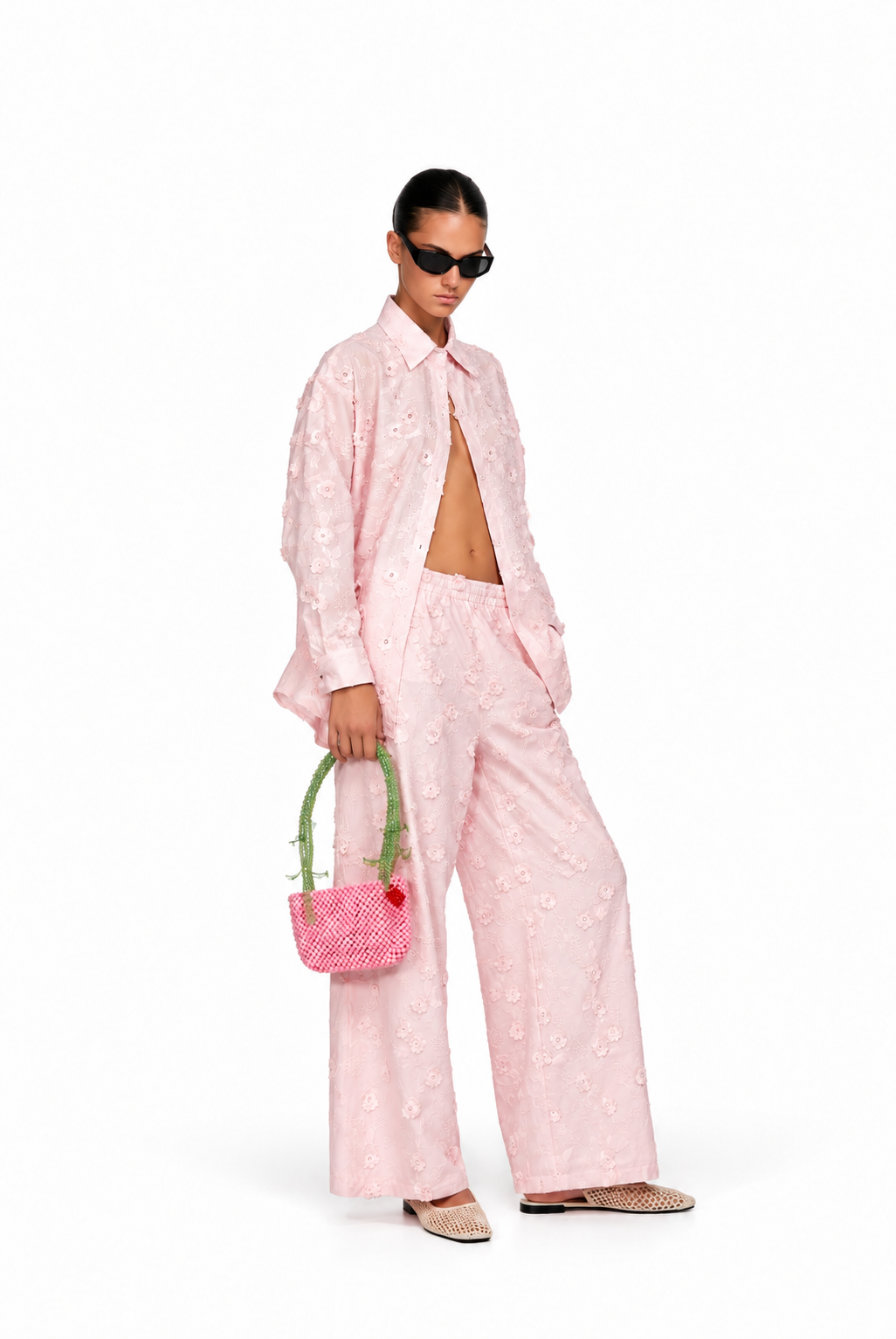 A model wearing a light pink embroidered two-piece set consisting of a long-sleeve button-up shirt and matching wide-leg trousers, styled with a small pink patterned handbag, sunglasses, and beige flat shoes against a white background.