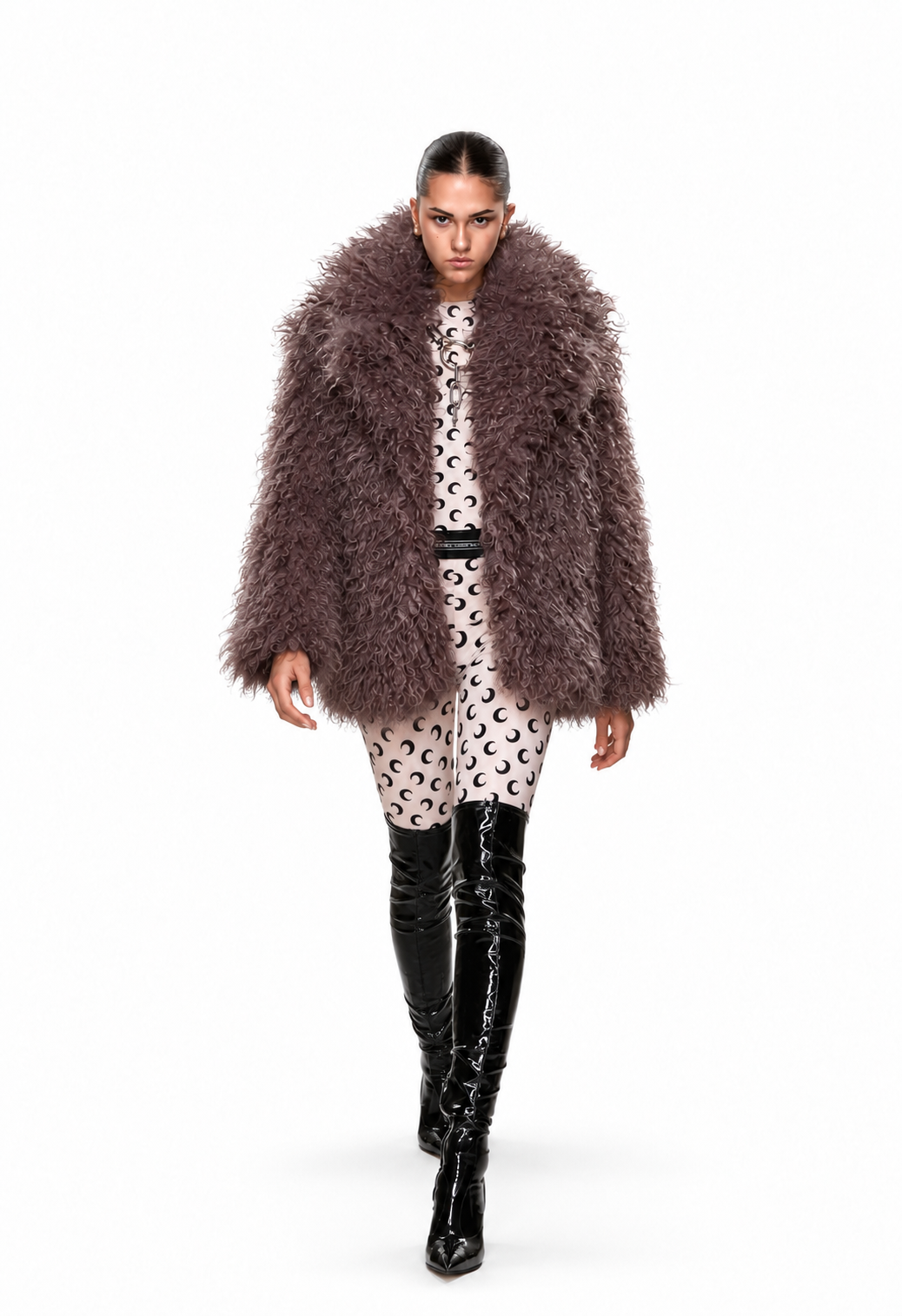 A model walks against a white studio background wearing a shaggy, oversized brown faux-fur coat over a beige and black patterned dress, paired with black patent leather over-the-knee boots.