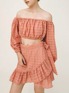 A model wearing a dusty rose two-piece set featuring an off-the-shoulder long-sleeve crop top and a matching ruffled mini skirt with a side tie, both made of a dotted eyelet fabric.