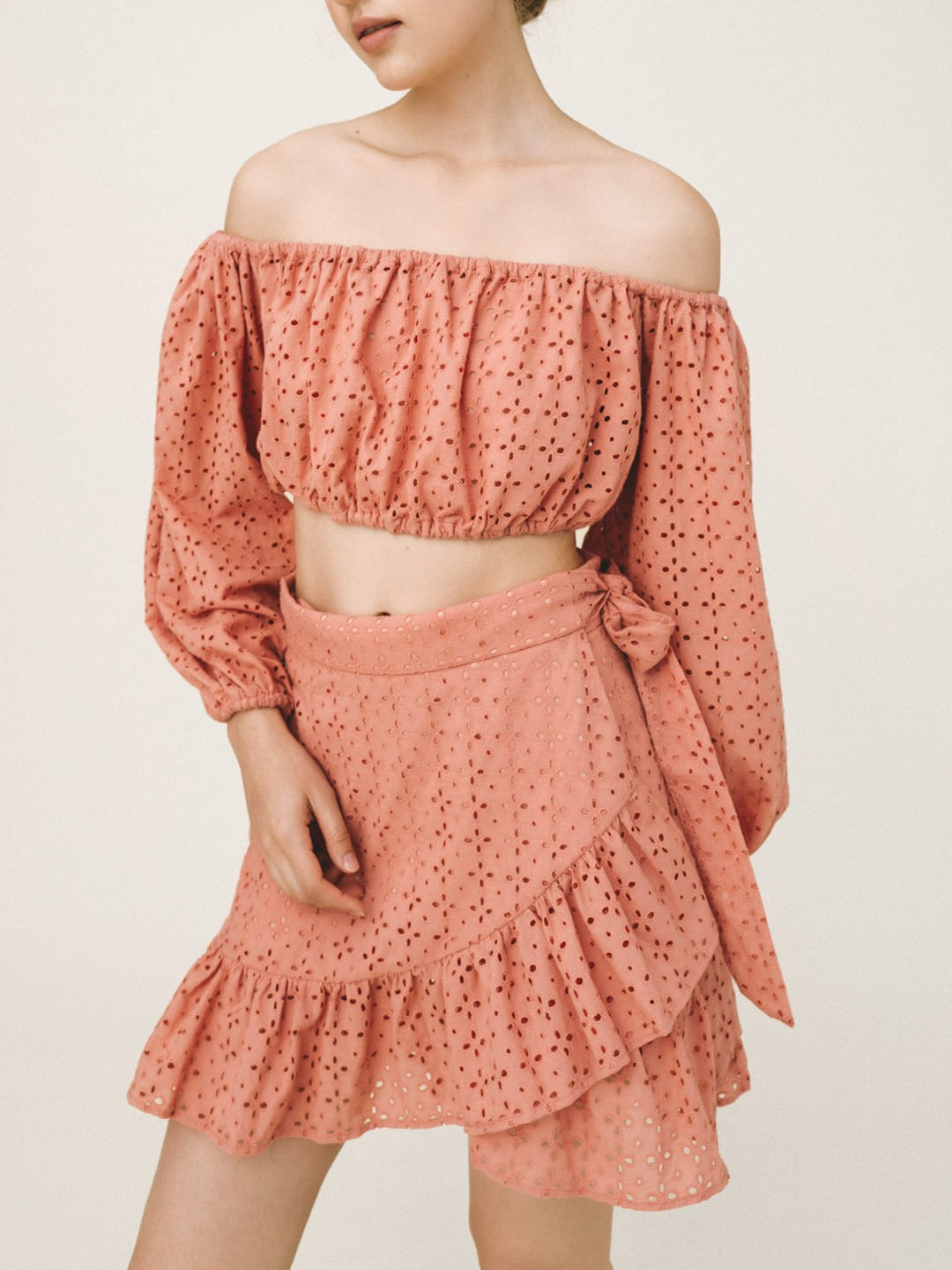 A model wearing a dusty rose two-piece set featuring an off-the-shoulder long-sleeve crop top and a matching ruffled mini skirt with a side tie, both made of a dotted eyelet fabric.