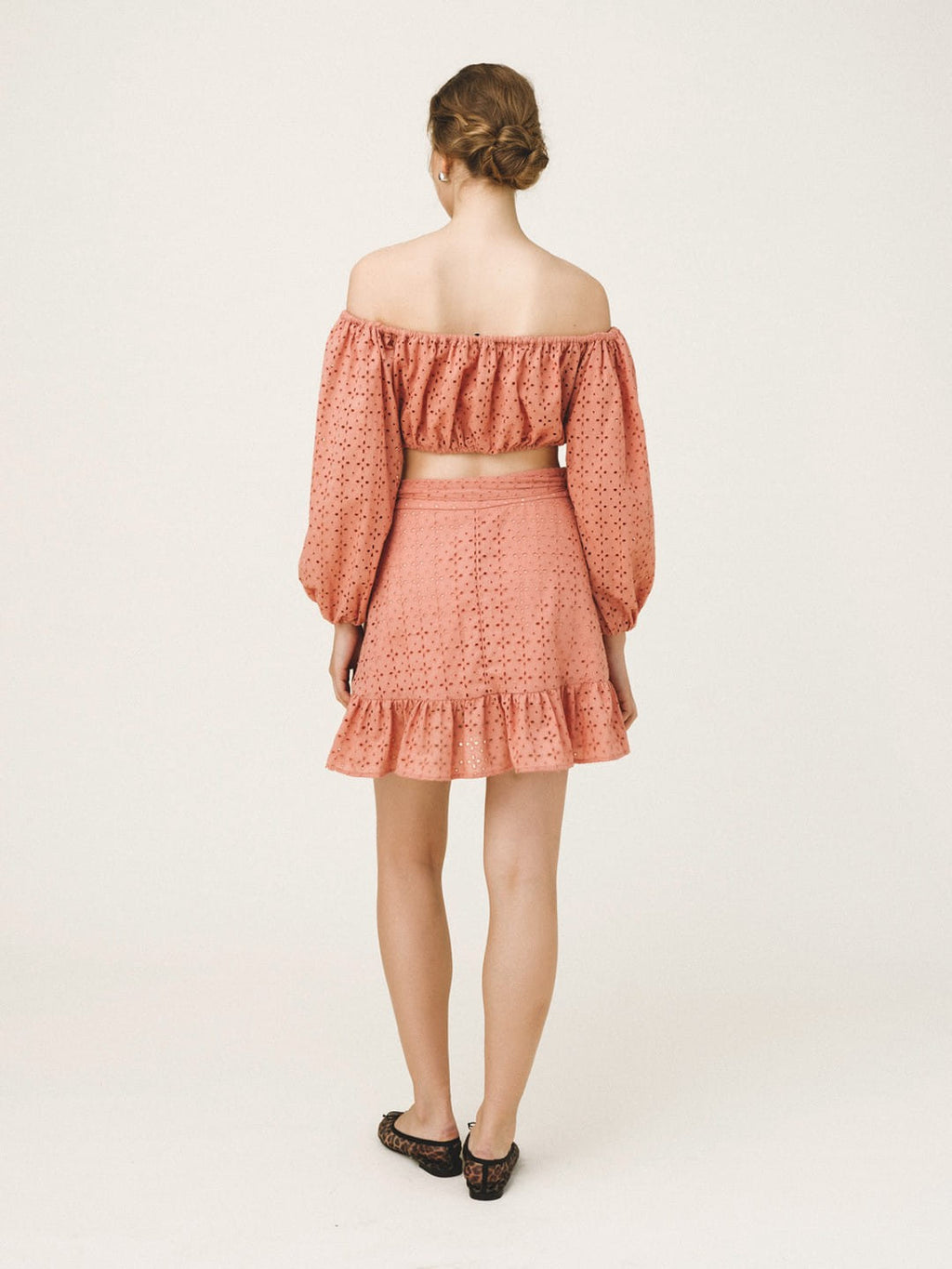 A woman viewed from the back wearing a matching two-piece set consisting of an off-the-shoulder long-sleeve crop top and a ruffled mini skirt, both in a dusty coral pink color with a textured eyelet pattern. She is wearing black flat shoes against a plain off-white studio background.