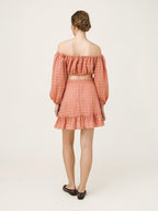 A woman viewed from the back wearing a matching two-piece set consisting of an off-the-shoulder long-sleeve crop top and a ruffled mini skirt, both in a dusty coral pink color with a textured eyelet pattern. She is wearing black flat shoes against a plain off-white studio background.