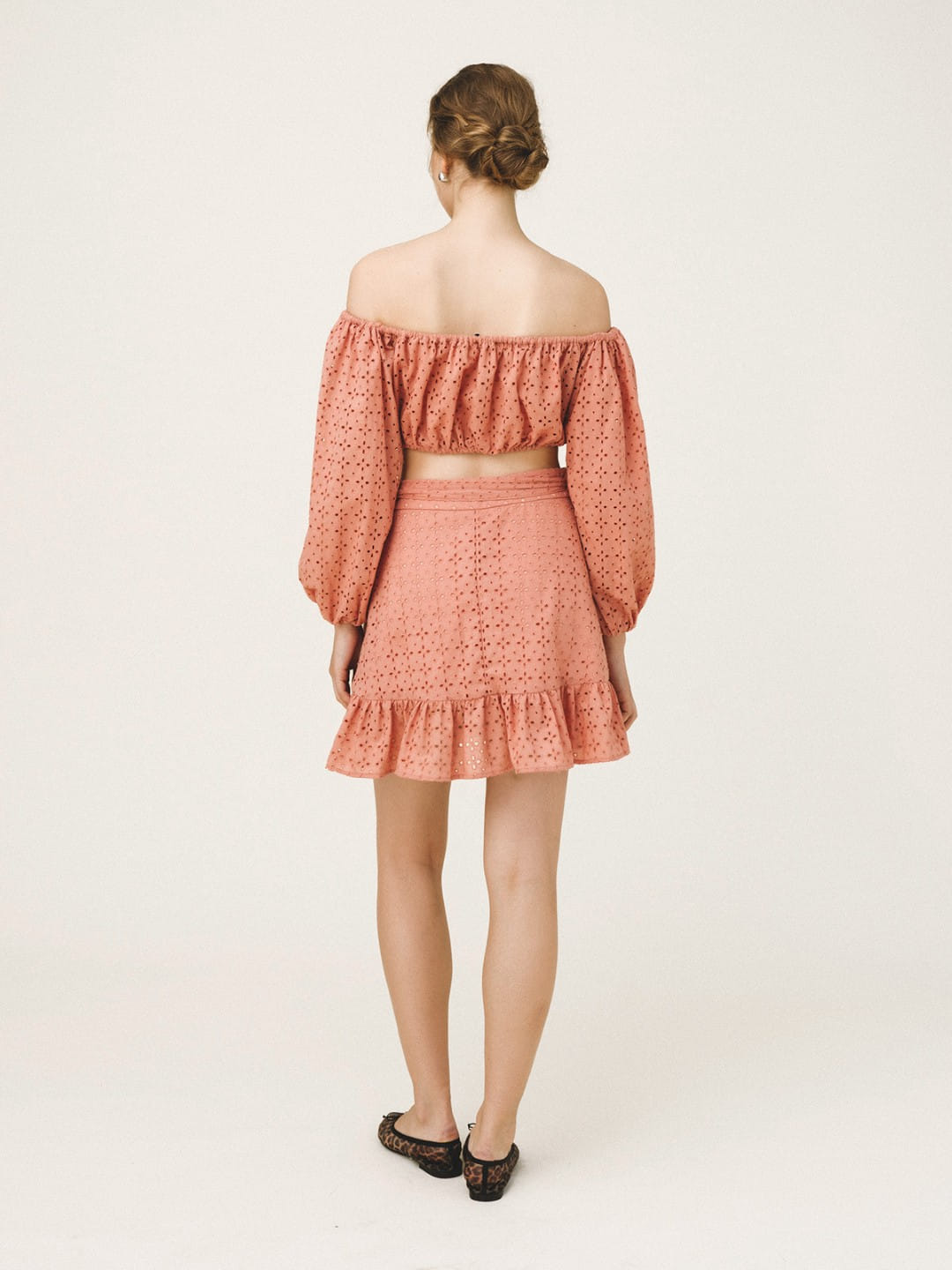 A woman viewed from the back wearing a matching two-piece set consisting of an off-the-shoulder long-sleeve crop top and a ruffled mini skirt, both in a dusty coral pink color with a textured eyelet pattern. She is wearing black flat shoes against a plain off-white studio background.