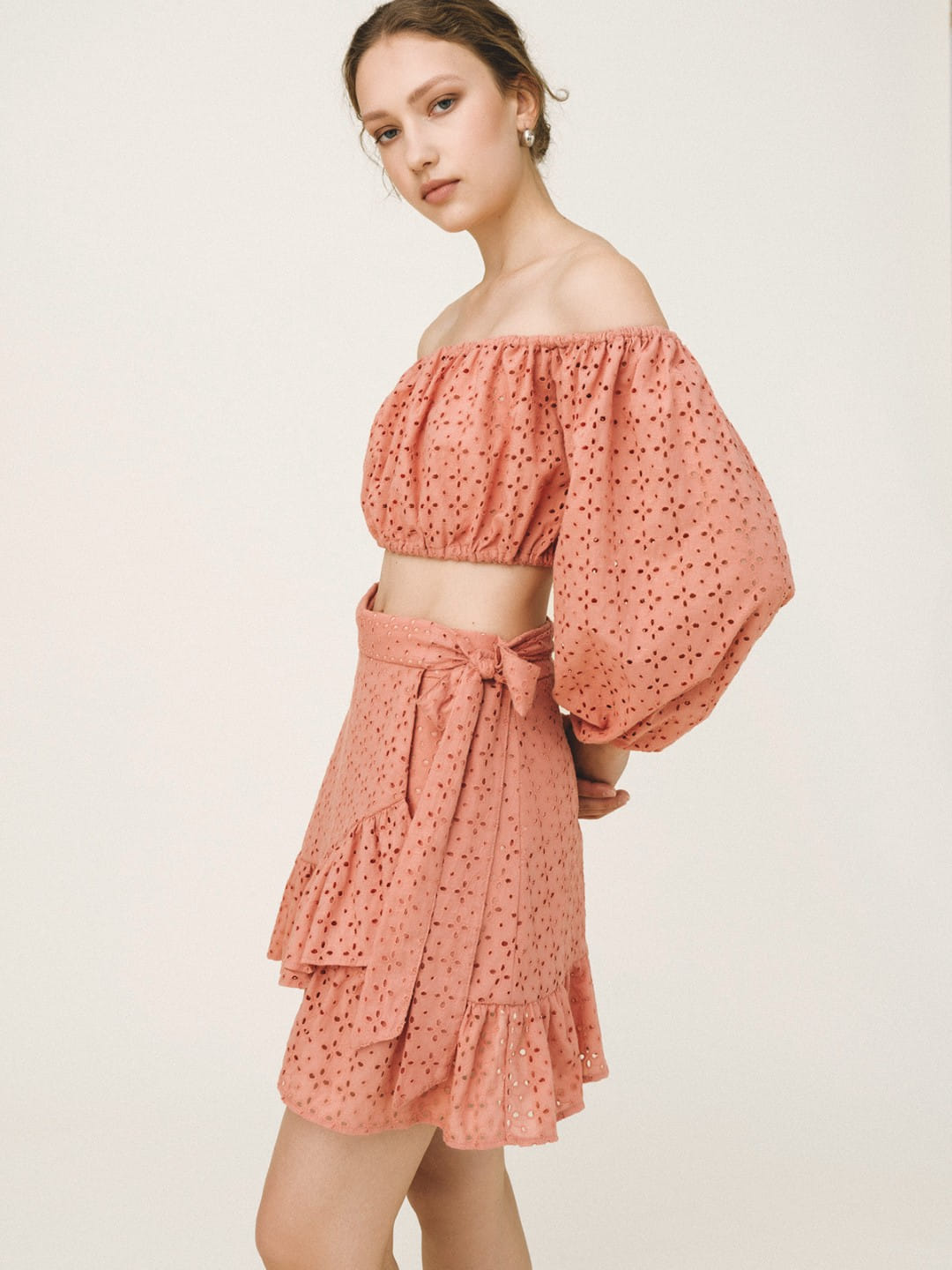 A woman modeling a two-piece dusty rose eyelet set consisting of an off-the-shoulder crop top with balloon sleeves and a matching tiered ruffled mini skirt with a tie waist, against a plain light background.