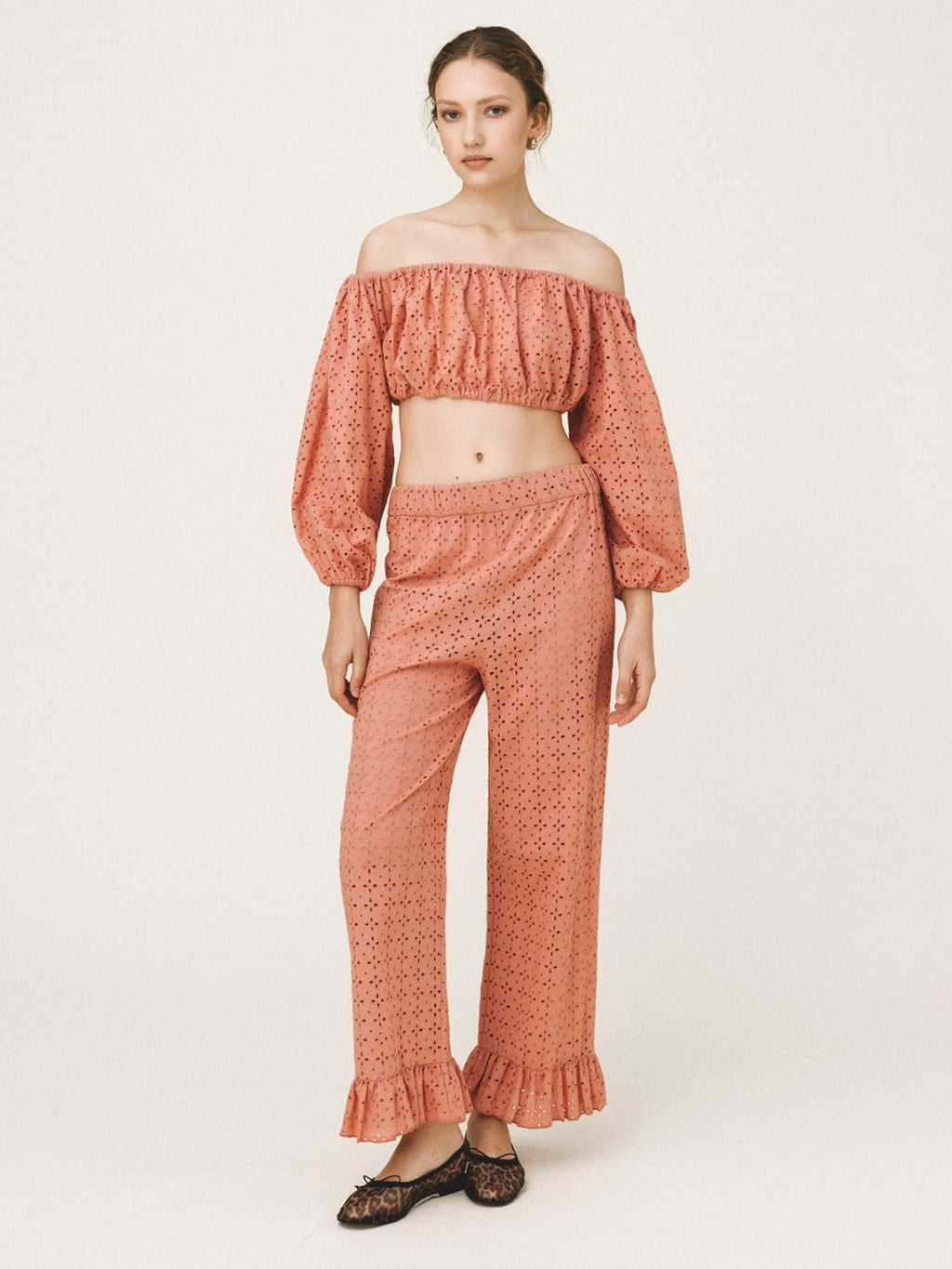 A model wearing a matching terracotta-colored two-piece set consisting of an off-the-shoulder long-sleeve crop top and high-waisted wide-leg pants with ruffled hems, paired with black lace-up flats, against a plain light beige studio background.