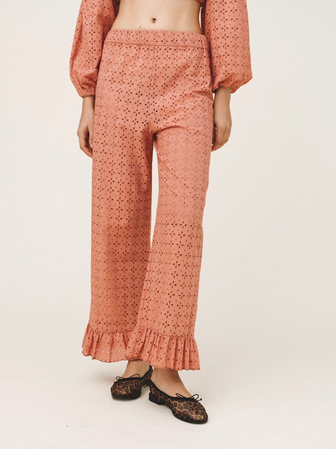 A model is shown from the waist down wearing peach-colored eyelet lace wide-leg pants with a ruffled hem and matching long-sleeve top, paired with black lace flat shoes, against a plain light background.