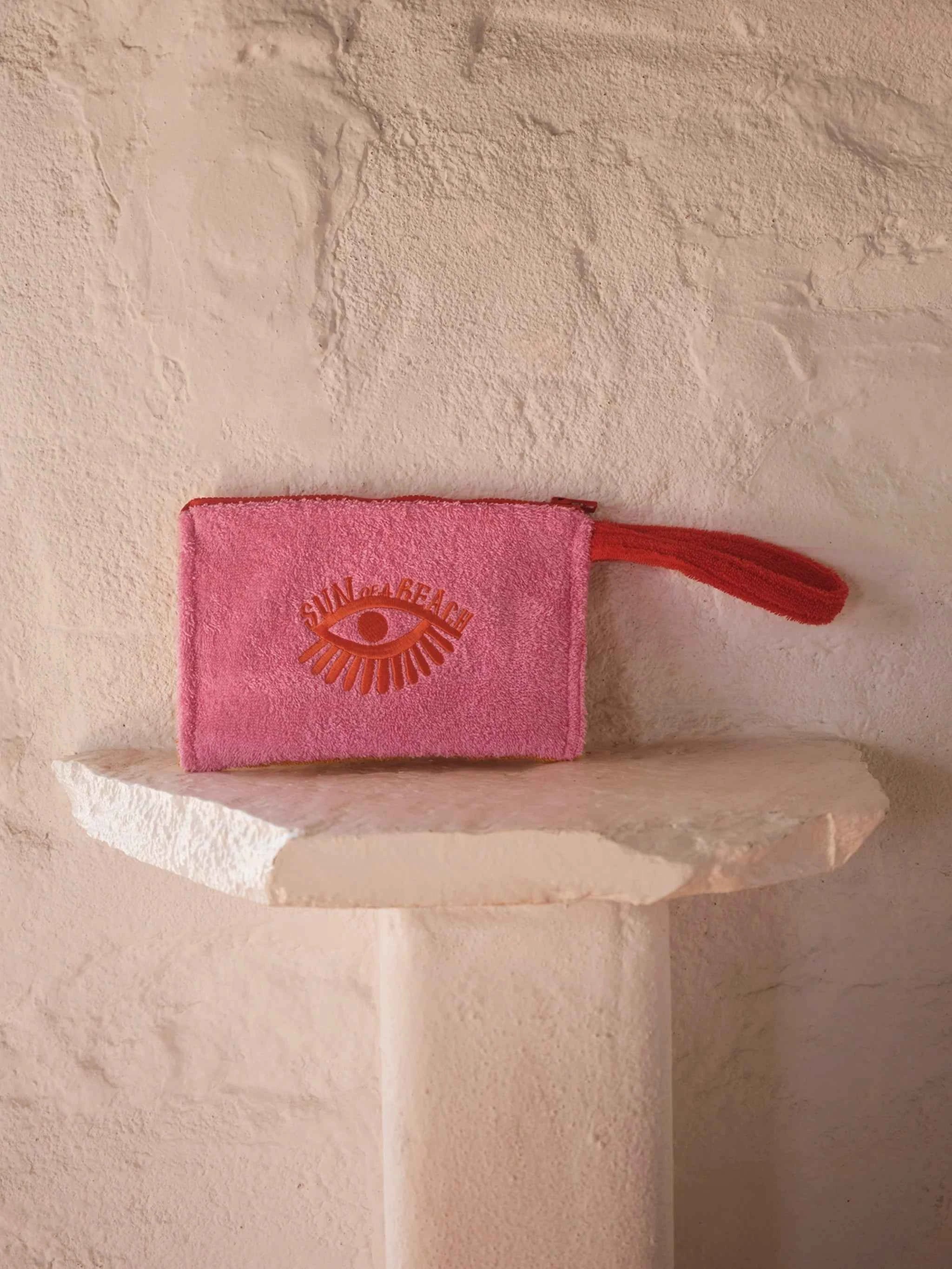 Pink pouch with a red handle on a white pedestal against a textured beige wall.