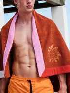 Man wearing a brown towel with pink trim and 'Sun Bum' logo, standing outdoors.