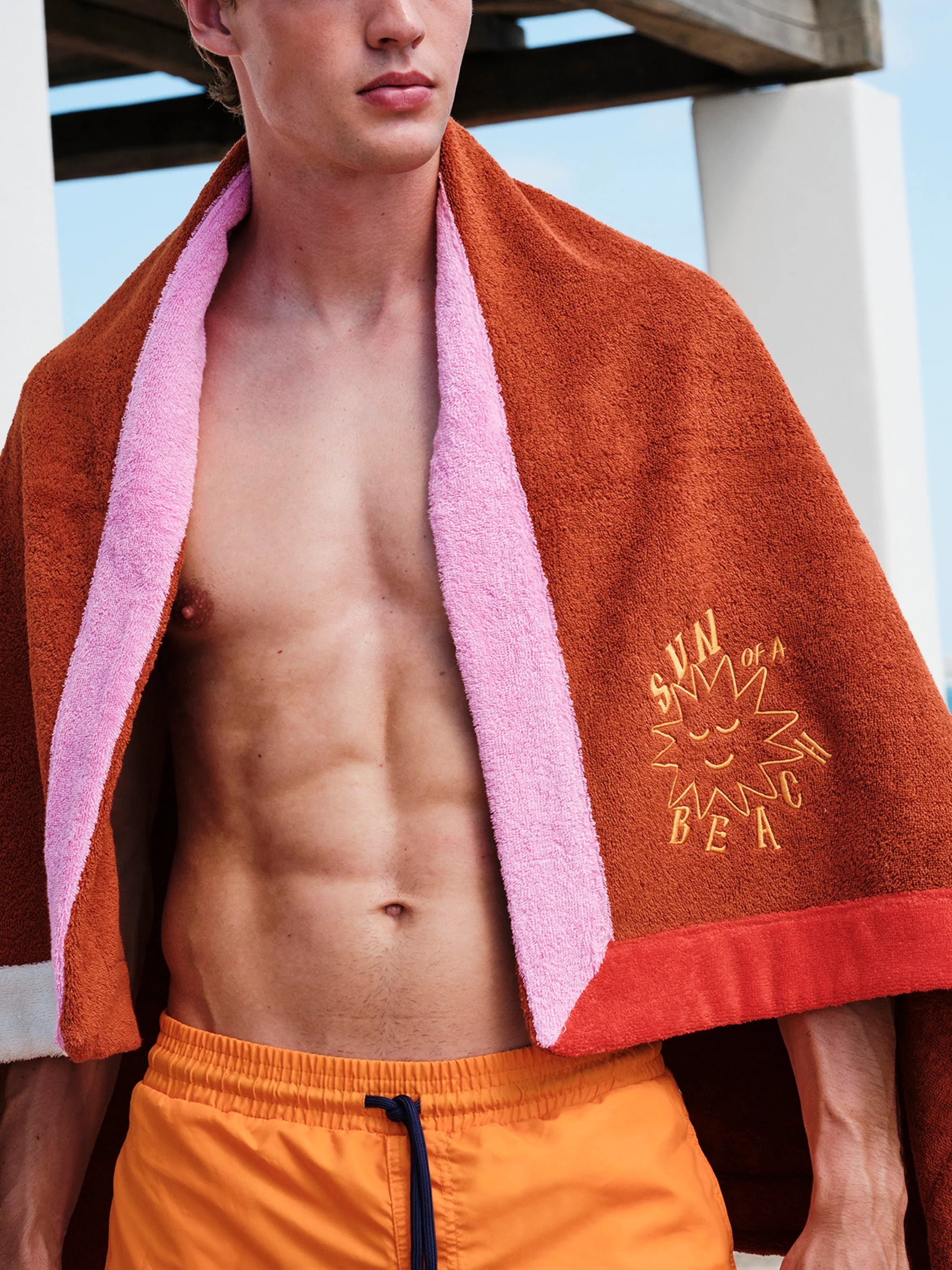 Man wearing a brown towel with pink trim and 'Sun Bum' logo, standing outdoors.
