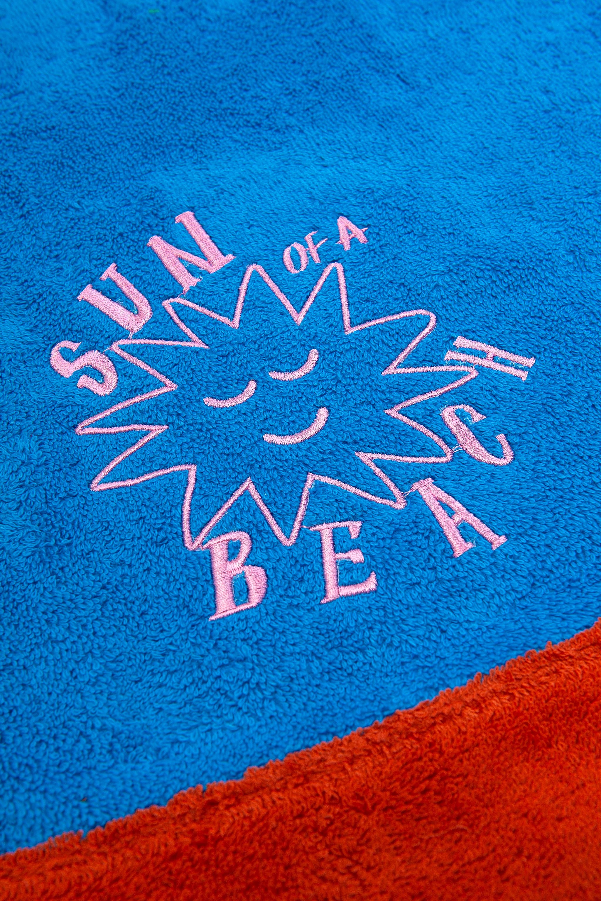 Blue towel with pink text and sun design, featuring the phrase 'Sun of a Beach'.
