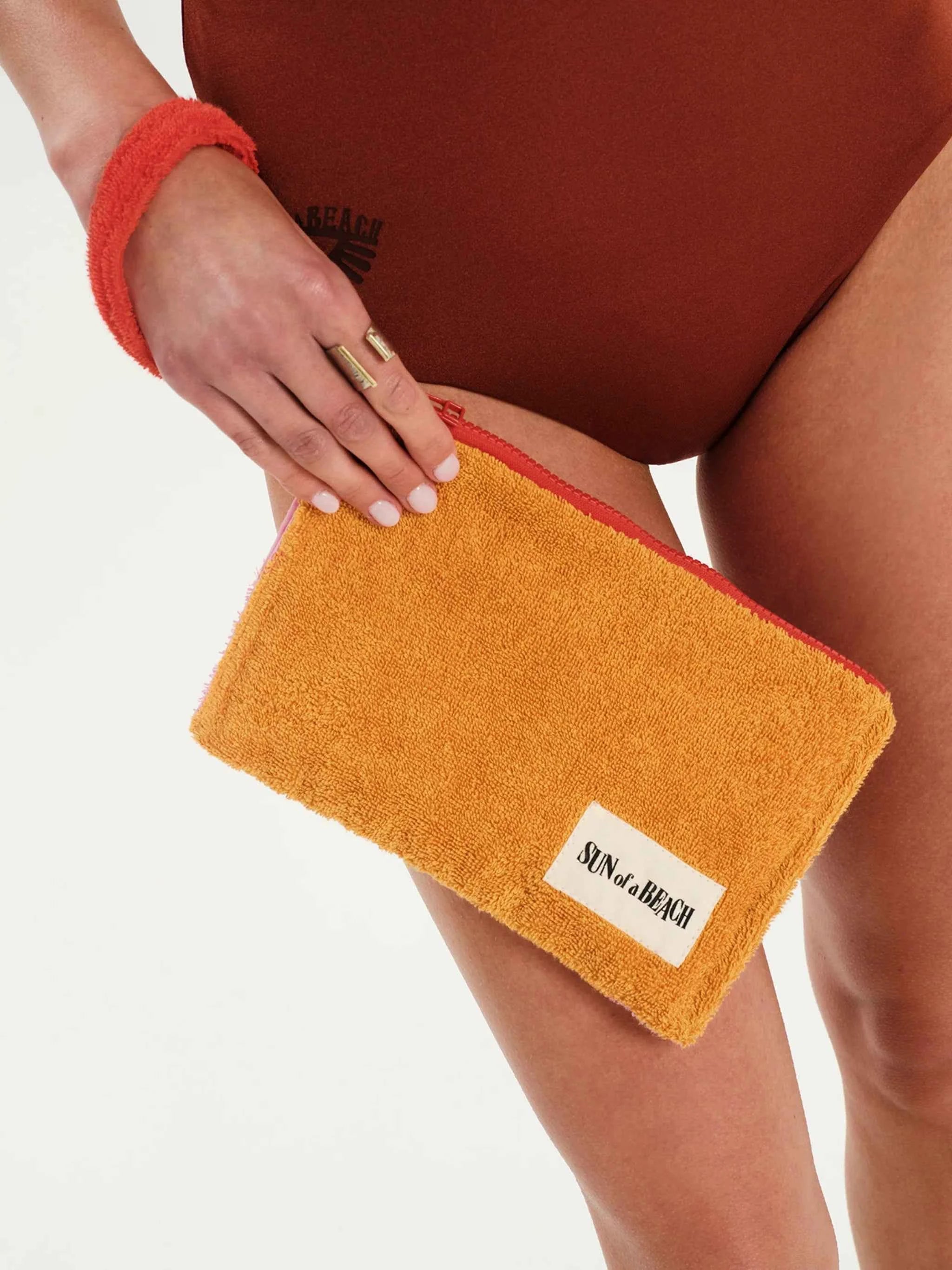 Person holding a mustard yellow pouch with a brand label on a neutral background