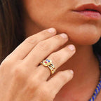 Close-up of a hand wearing a gold ring with colorful gemstones, blurred background