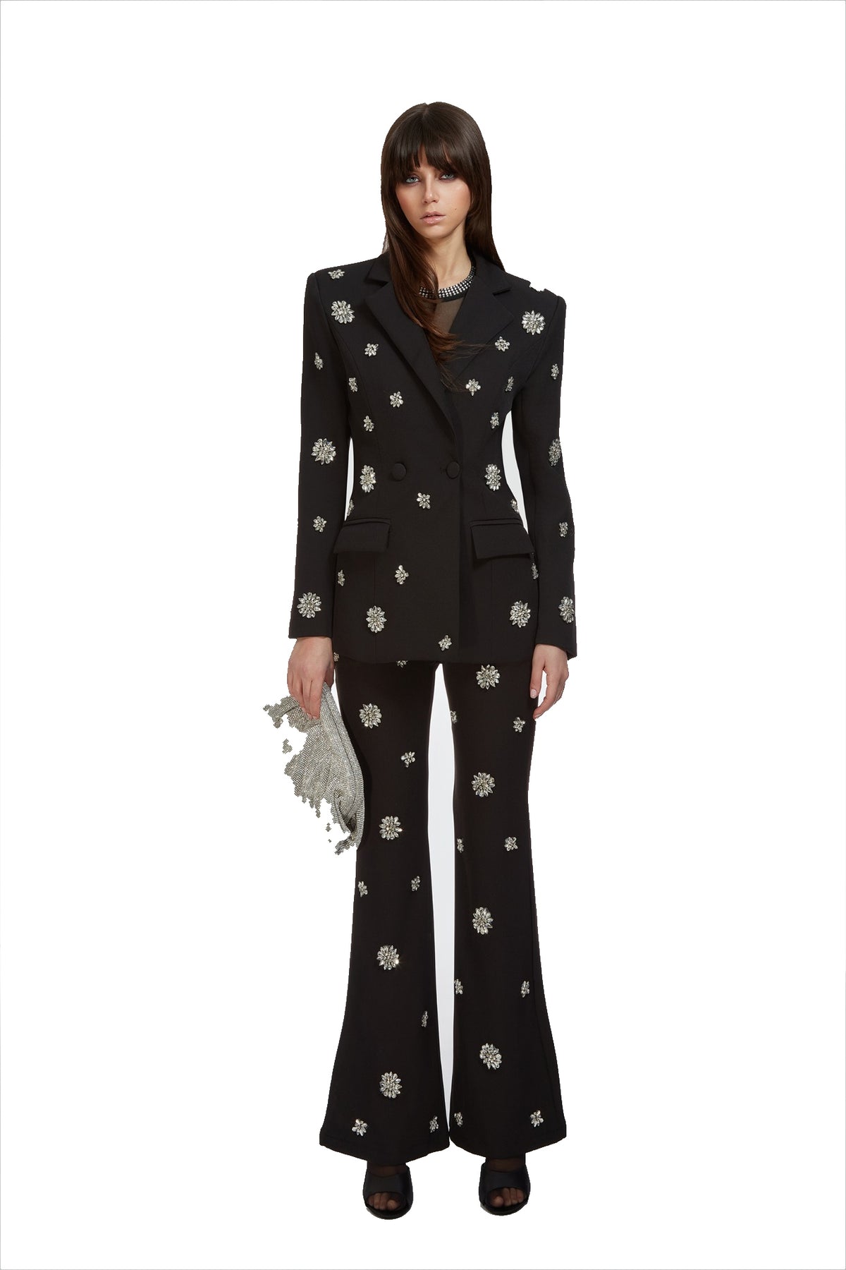 A model stands against a white background wearing a black double-breasted blazer and matching flared trousers, both decorated with small white floral patterns. She is holding a small silver clutch.