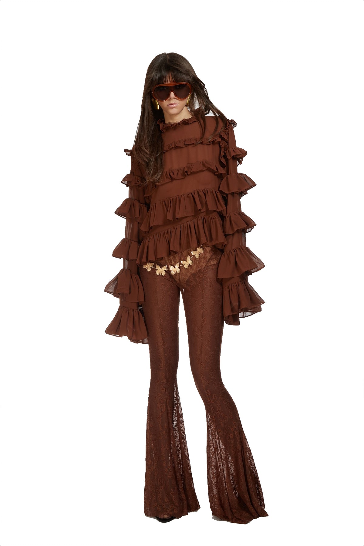 A model wearing a brown bohemian-style two-piece set consisting of a long-sleeve top with dramatic ruffles and matching flared pants with lace inserts, accessorized with sunglasses and a flower belt.