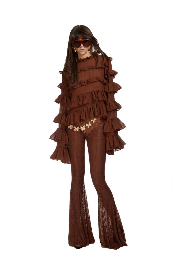 A model wearing a brown bohemian-style two-piece set consisting of a long-sleeve top with dramatic ruffles and matching flared pants with lace inserts, accessorized with sunglasses and a flower belt.