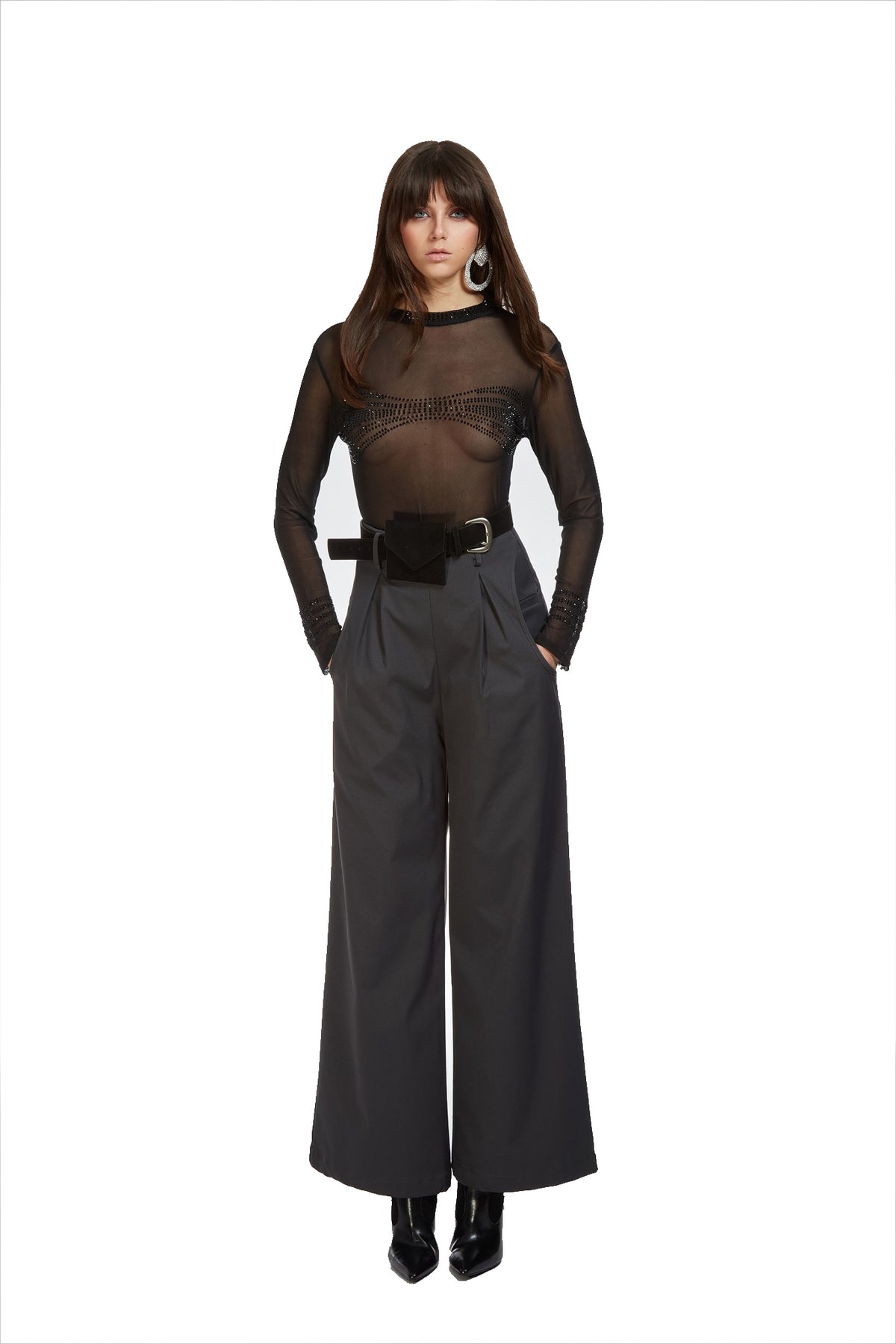 A model wearing a sheer black long-sleeve top with a black lace-trimmed neckline, paired with high-waisted dark gray wide-leg trousers, a black belt, and black boots, set against a plain white background.