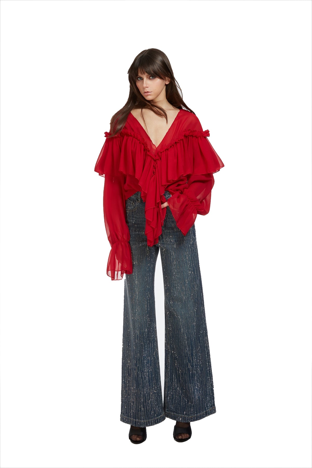 A model wearing a vibrant red off-the-shoulder blouse with dramatic ruffles and long sheer sleeves, paired with wide-leg blue jeans and dark brown shoes, set against a plain white background.