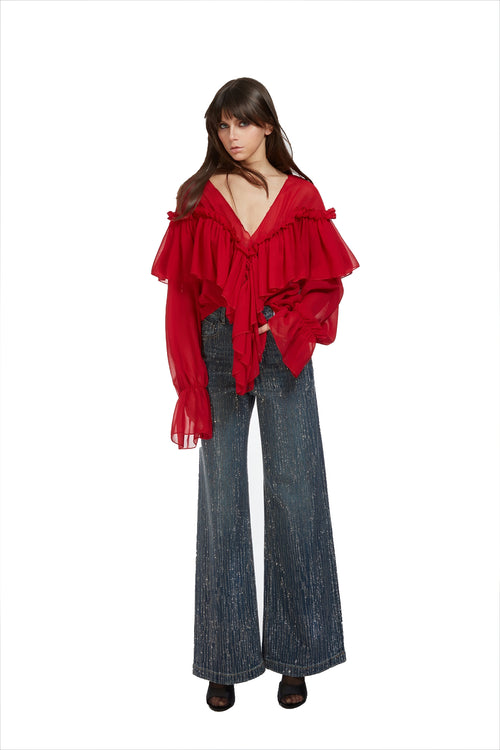 A model wearing a vibrant red off-the-shoulder blouse with dramatic ruffles and long sheer sleeves, paired with wide-leg blue jeans and dark brown shoes, set against a plain white background.