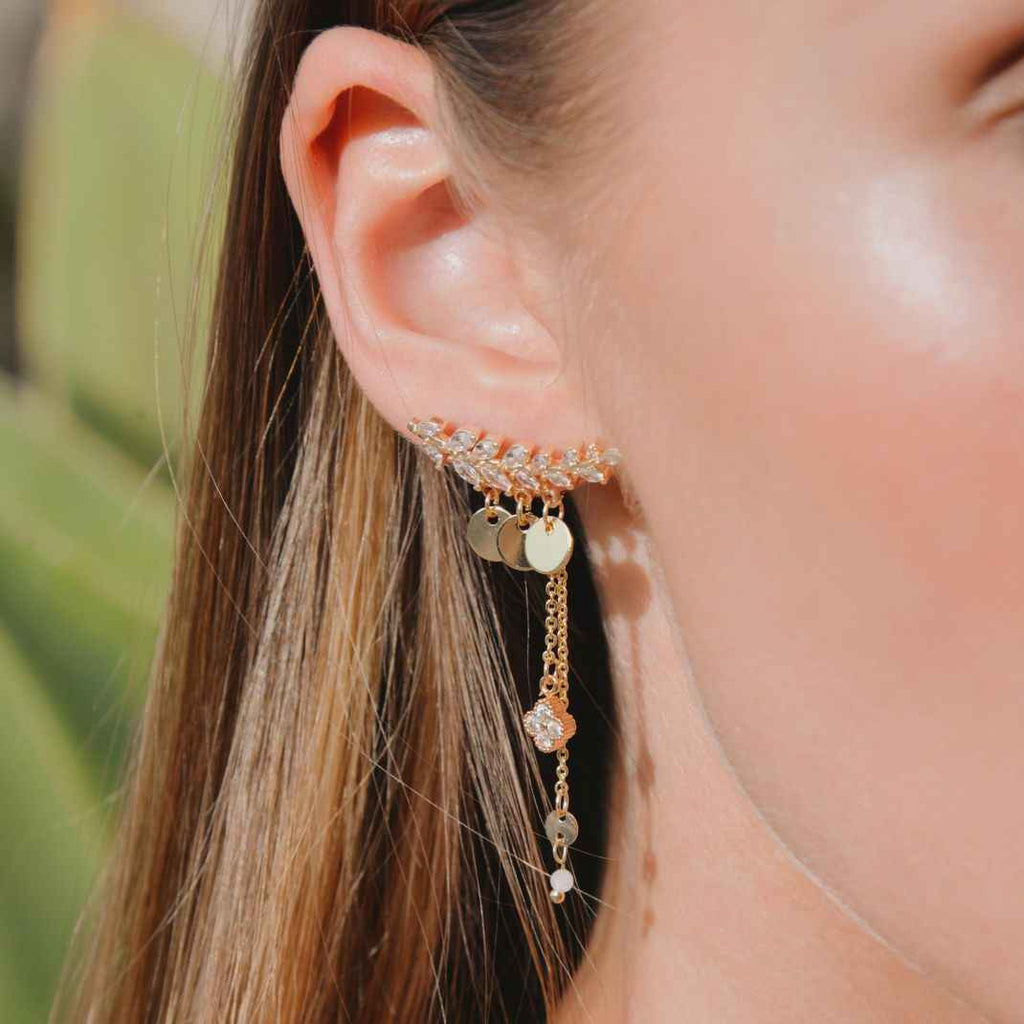 Close-up of a person wearing a gold earring with dangling charms against a blurred background.