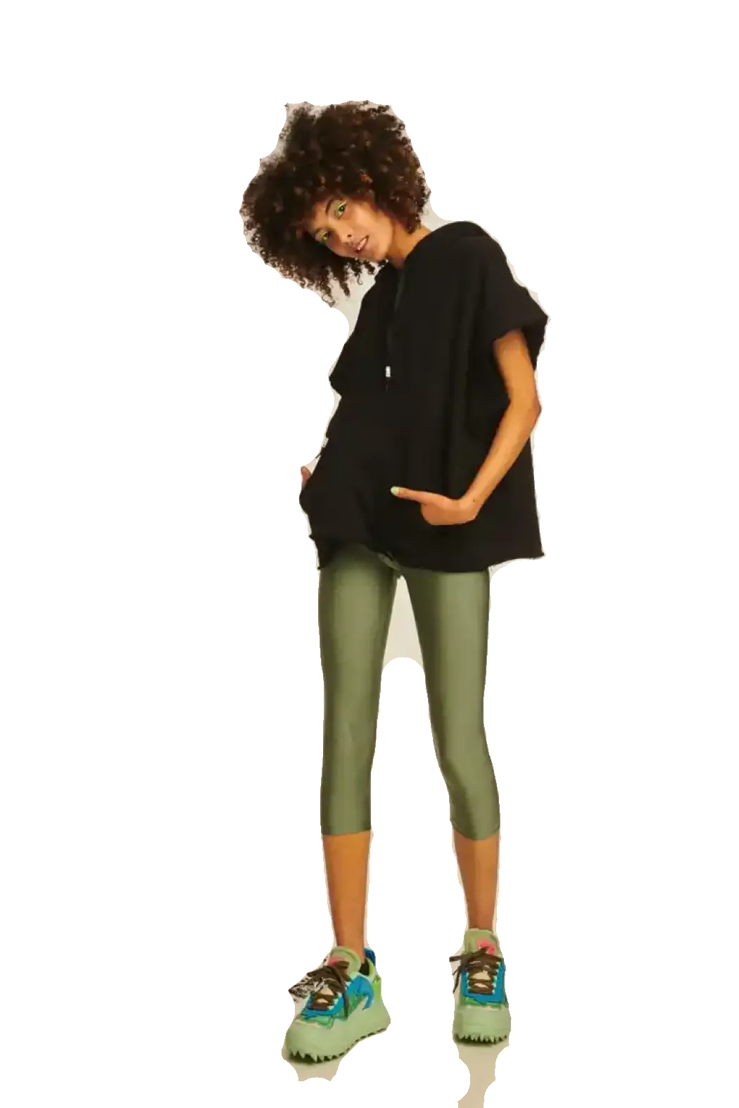 A model with an afro hairstyle poses in a black oversized t-shirt, olive green capri leggings, and colorful sneakers against a solid white background.