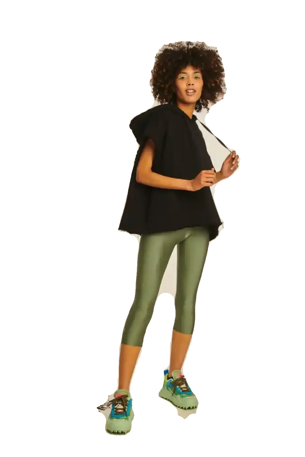 A woman with a large curly afro stands in a studio wearing a black asymmetrical cape-style top, olive green leggings, and green sneakers with blue accents. She is holding a white sheet against a plain white background.